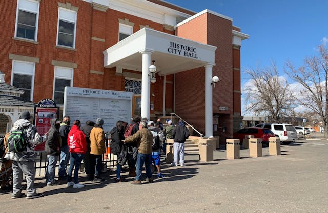A long line of people stand waiting for intake to the SWAP program outside a building with the title Historic City Hall. It is a bright day in winter, with no leaves on nearby trees.